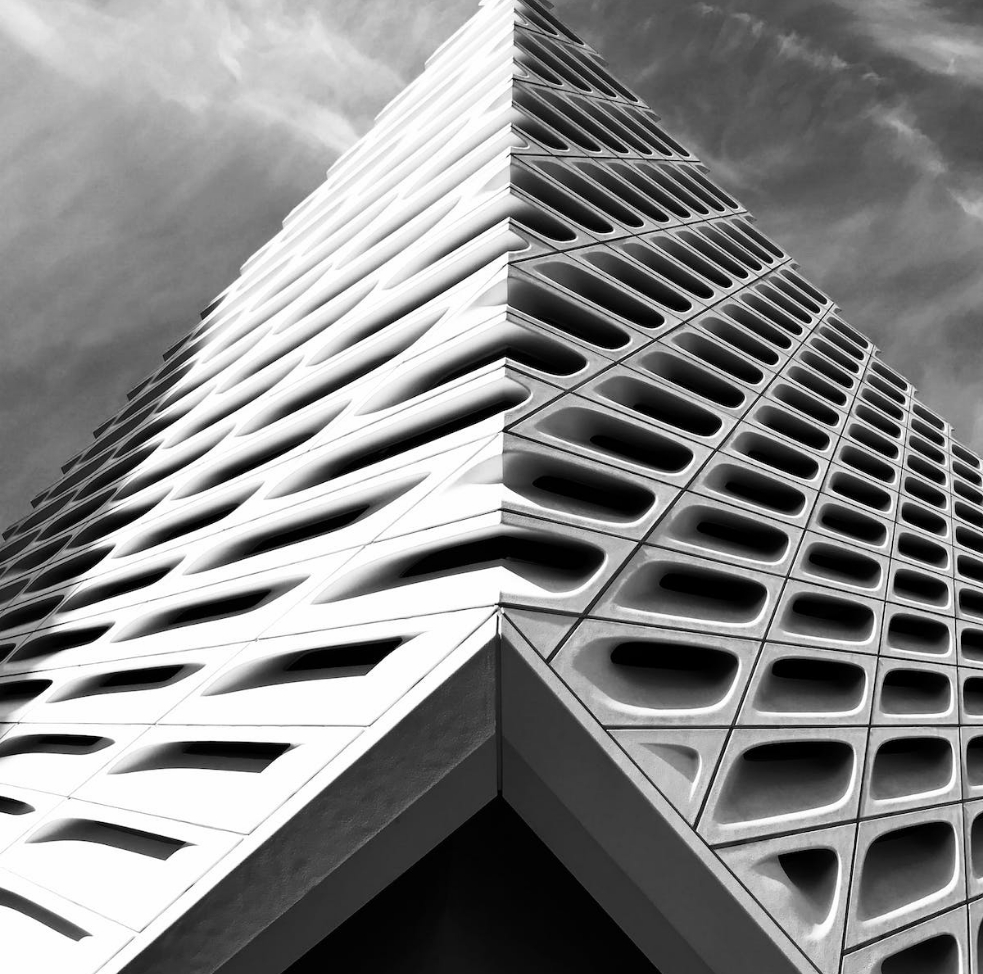 Black and white photo looking up at the corner of a modern building with geometric, perforated facade panels against a partly cloudy sky.