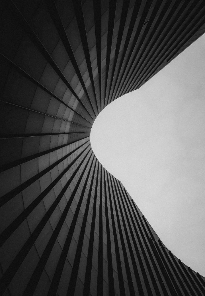 Low-angle black and white photo of a curved modern building with repetitive vertical lines converging to form an open space against a plain sky.