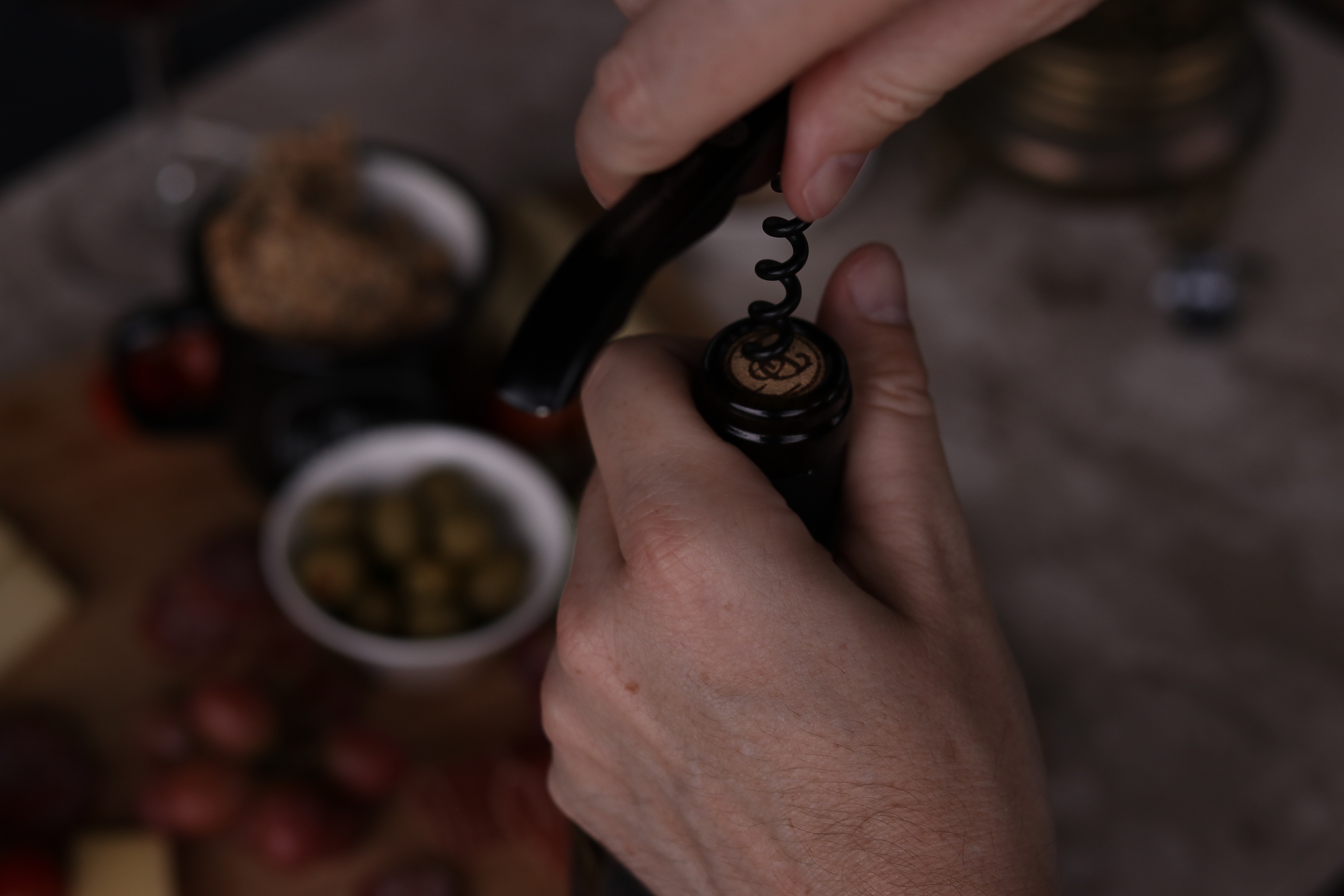 Hände die einen Korkenzieher verwenden, um eine Weinflasche zu öffnen, unscharfer Hintergrund mit Oliven und kalten Vorspeisen.