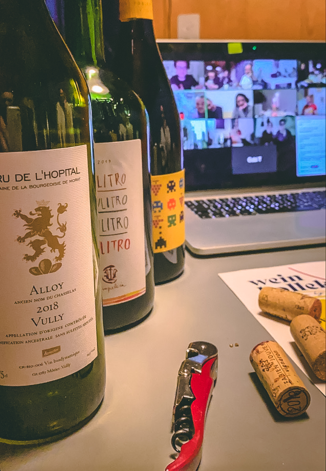 Drei Weinflaschen, ein Korkenzieher und mehrere Weinkorken auf einem Tisch vor einem Laptop mit Videokonferenz.