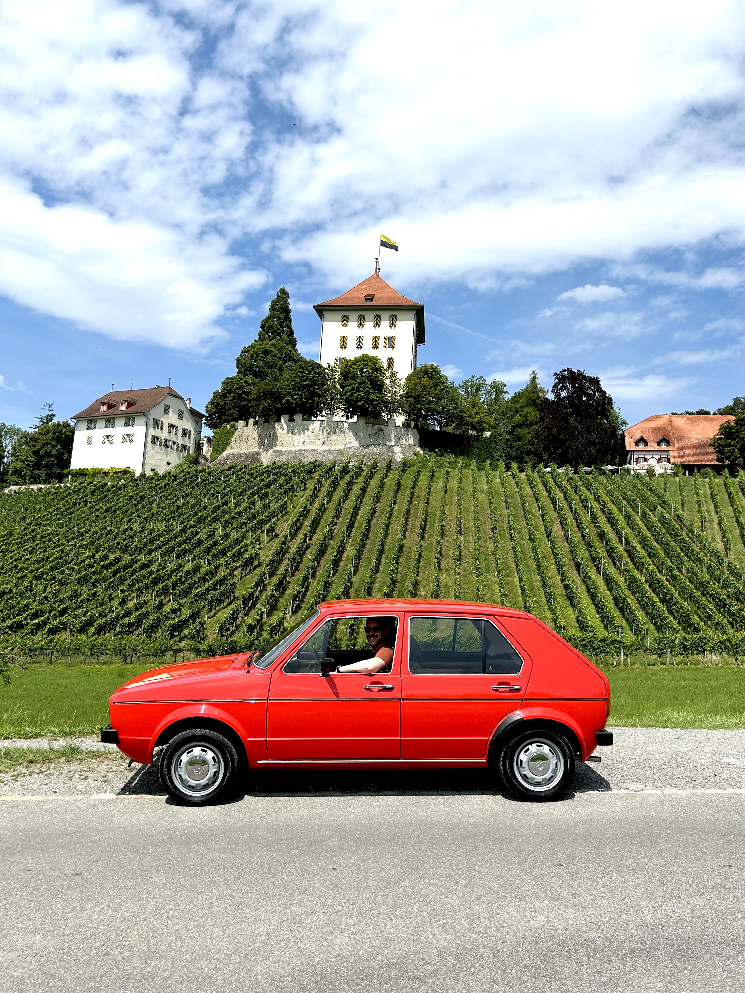 Roter Oldtimer-Pkw vor einer grünen Weinbergslage mit einem historischen Turm und Gebäuden im Hintergrund unter blauem Himmel.