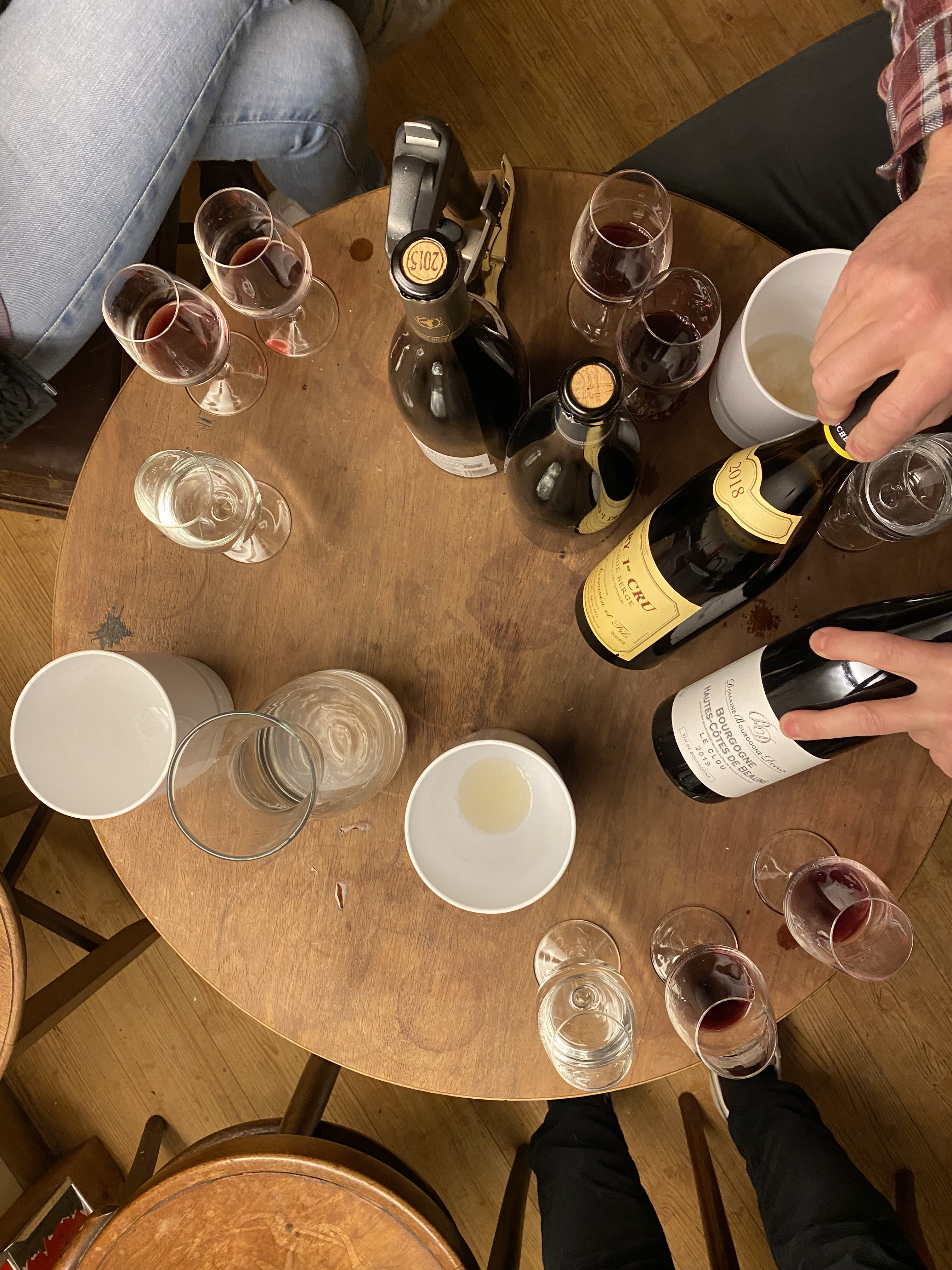 Holztisch mit mehreren Weinflaschen, Weingläsern mit Rot- und Weißwein und Personen, die Wein öffnen und trinken.