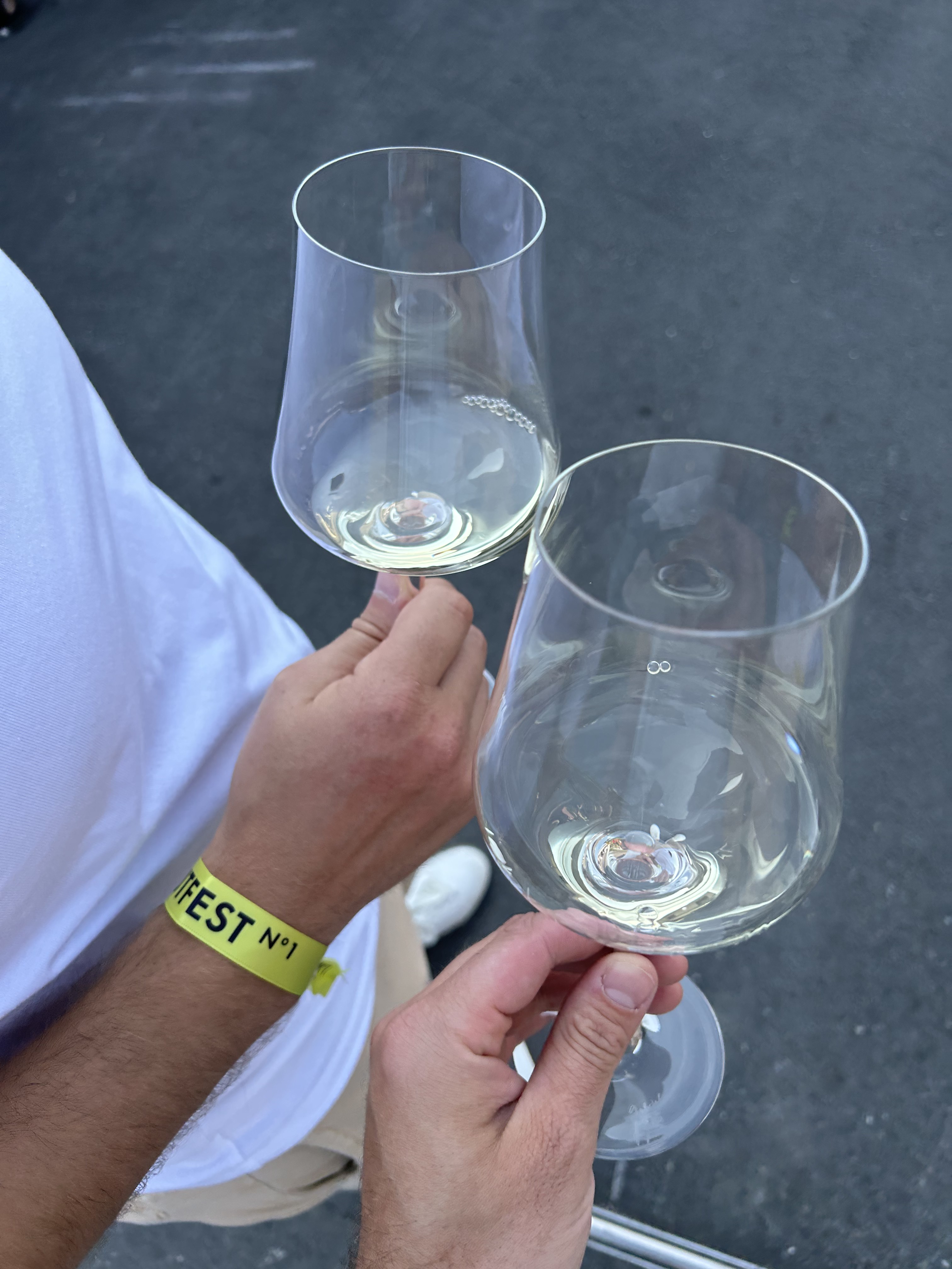 Zwei Personen halten transparente Weingläser mit weißem Wein, eine Person trägt ein gelbes Festival-Armband.
