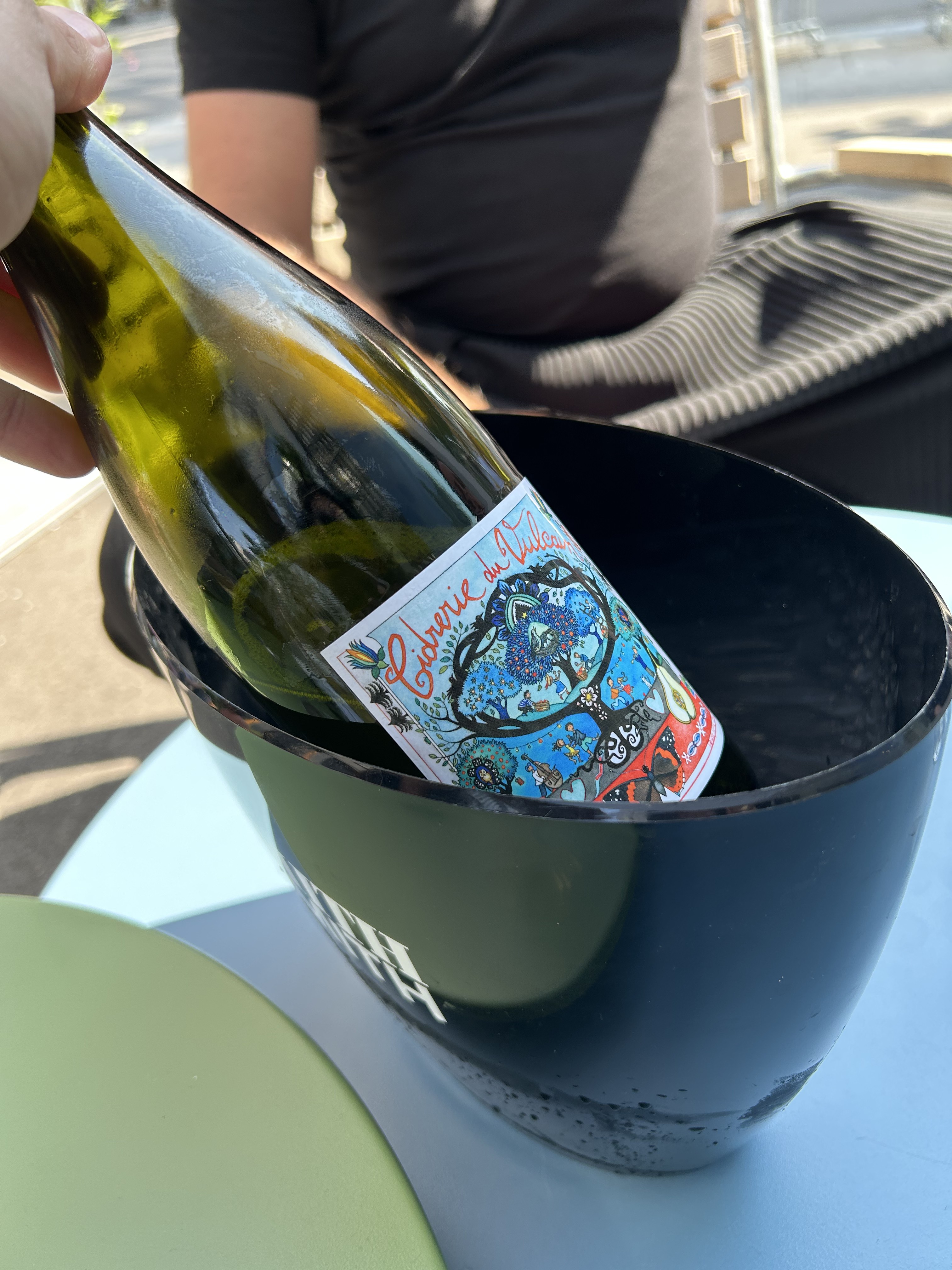 Hand hält eine Weinflasche mit einem bunten Etikett 'Cuvée du Vulcan' in einem schwarzen Eiskübel auf einem hellblauen Tisch.