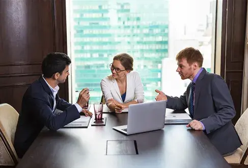 Three business professionals seated at a table, each using a laptop, engaged in a collaborative discussion.