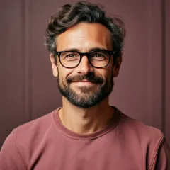 Mann mittleren Alters mit dunklem lockigem Haar, Bart und Brille lächelt freundlich, trägt ein rosafarbenes Oberteil, vor braunem Hintergrund.