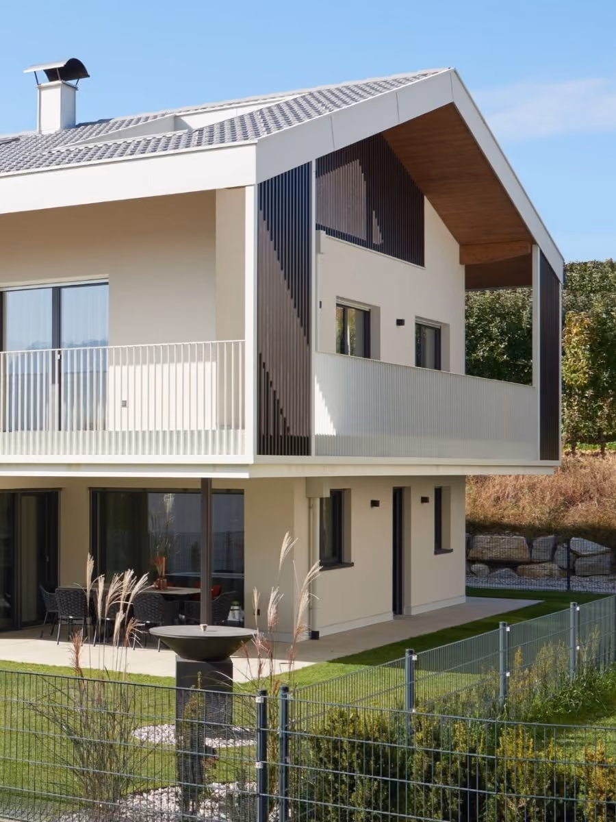 Moderne zweistöckige Wohnhaus mit großem Balkon auf der oberen Etage, weißer Fassade, verglasten Türen und Fenstern, einer Terrasse mit Sitzbereich im Erdgeschoss, umgeben von einem gepflegten Garten mit Zaun.