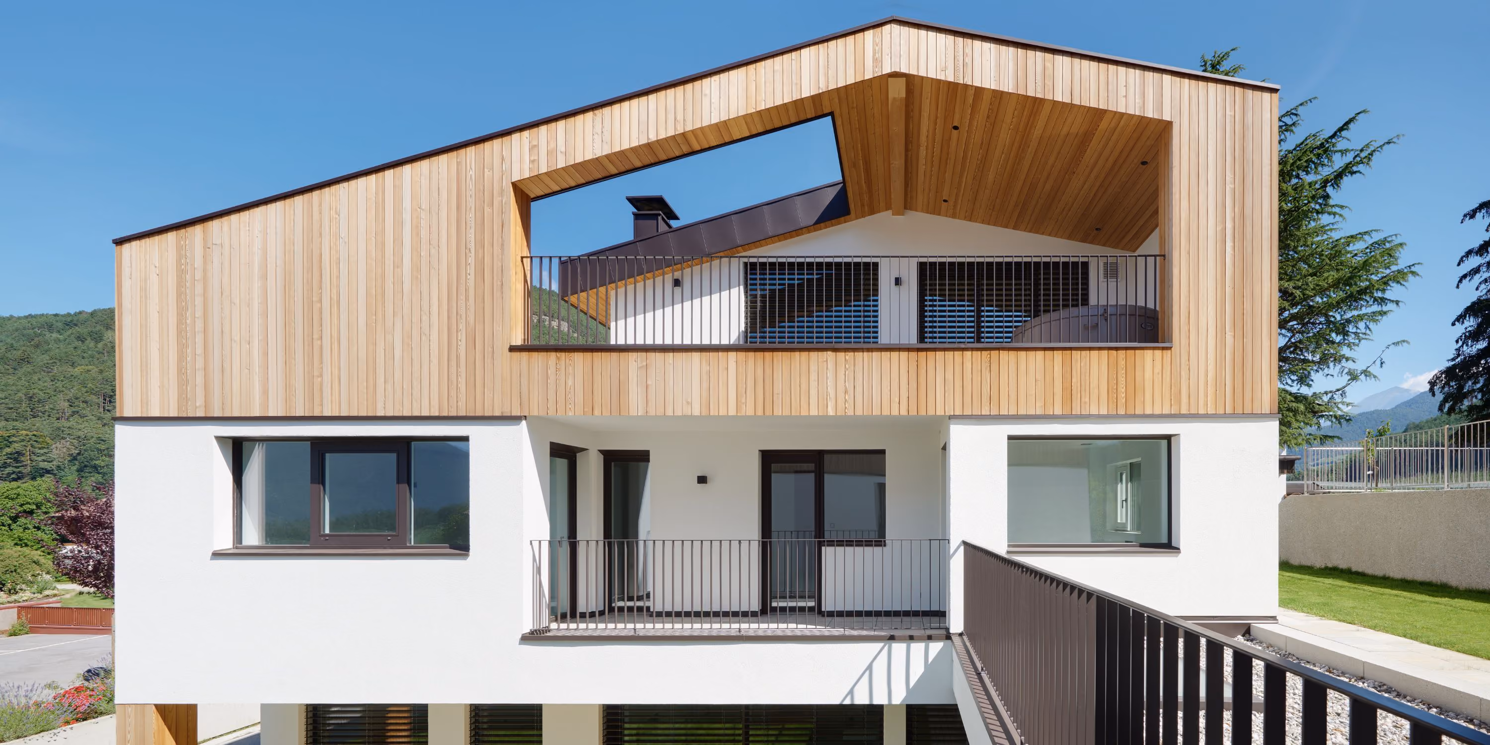 Modernes Wohnhaus mit weißer Fassade und Holzverkleidung im oberen Bereich, großem Balkon mit Metallgeländer und klar blauem Himmel im Hintergrund.