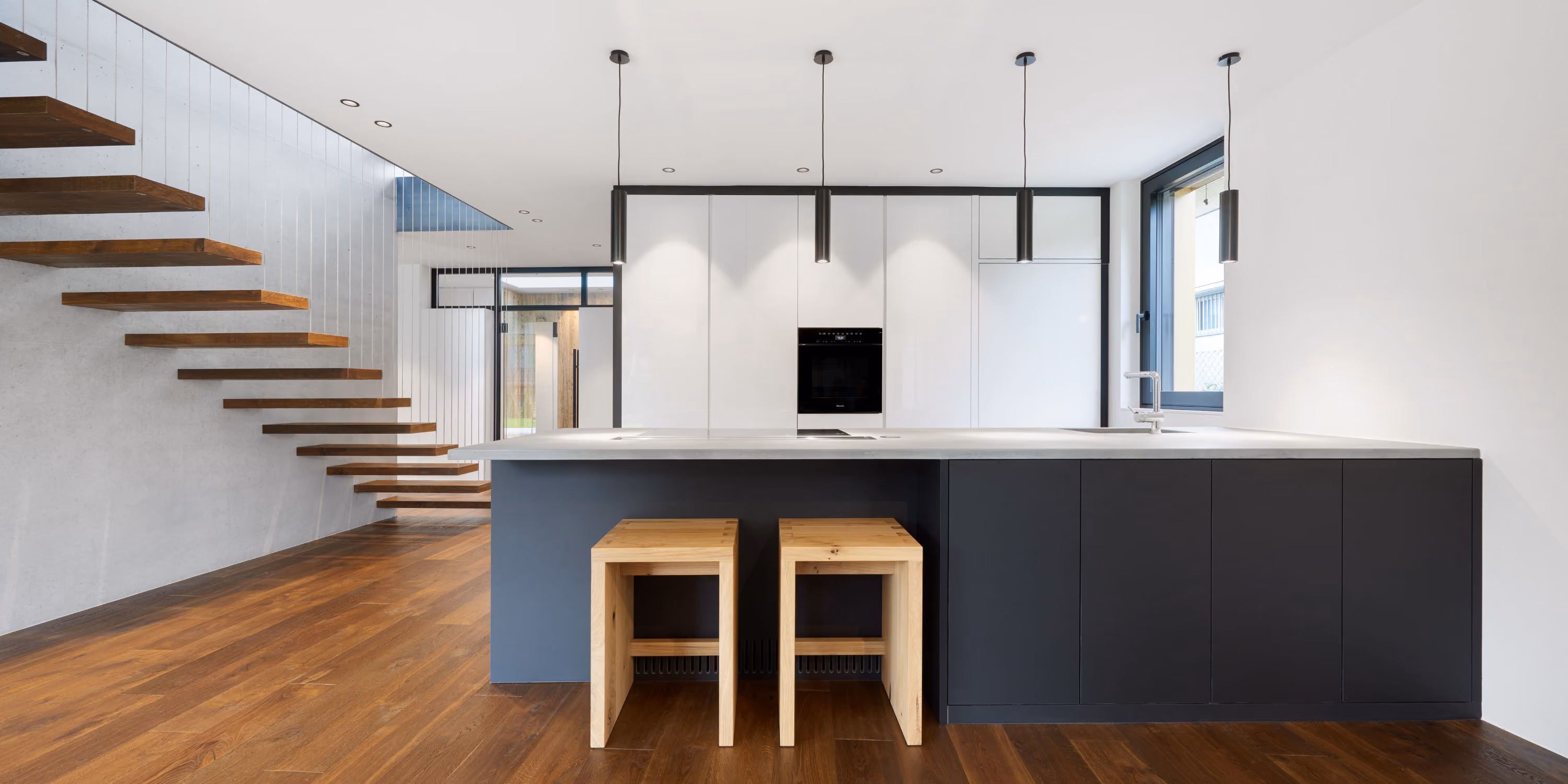 Moderne Küche mit hängenden schwarzen Pendelleuchten über Insel mit grauer Arbeitsplatte, zwei Holzhockern, schwarzen Unterschränken, weißen Hochschränken und schwebender Holztreppe an der Betonstrukturwand.