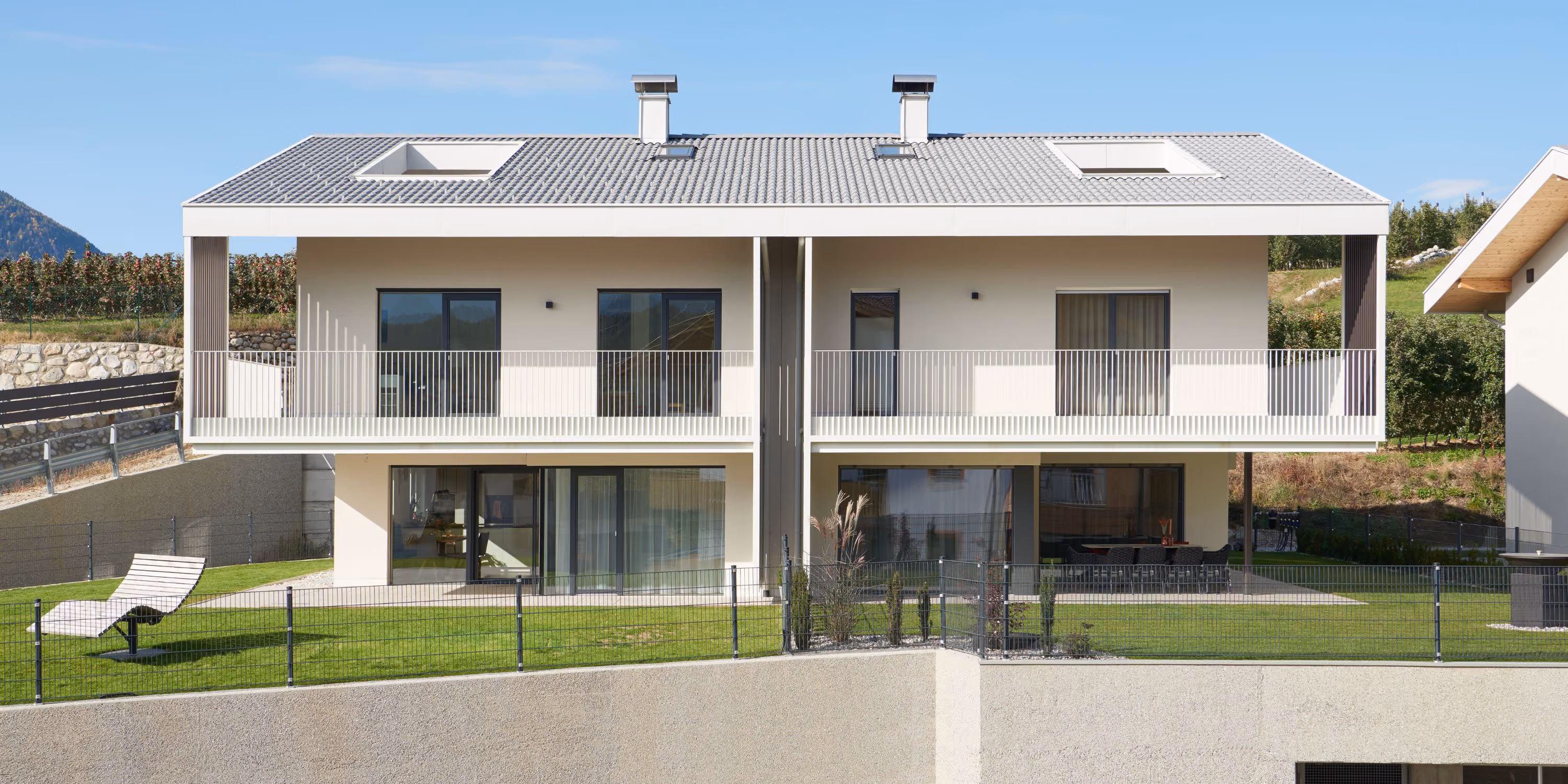 Modern duplex house with two floors and symmetrical design, featuring large windows, balconies on the upper floor, and a lawn with a sun lounger in front.