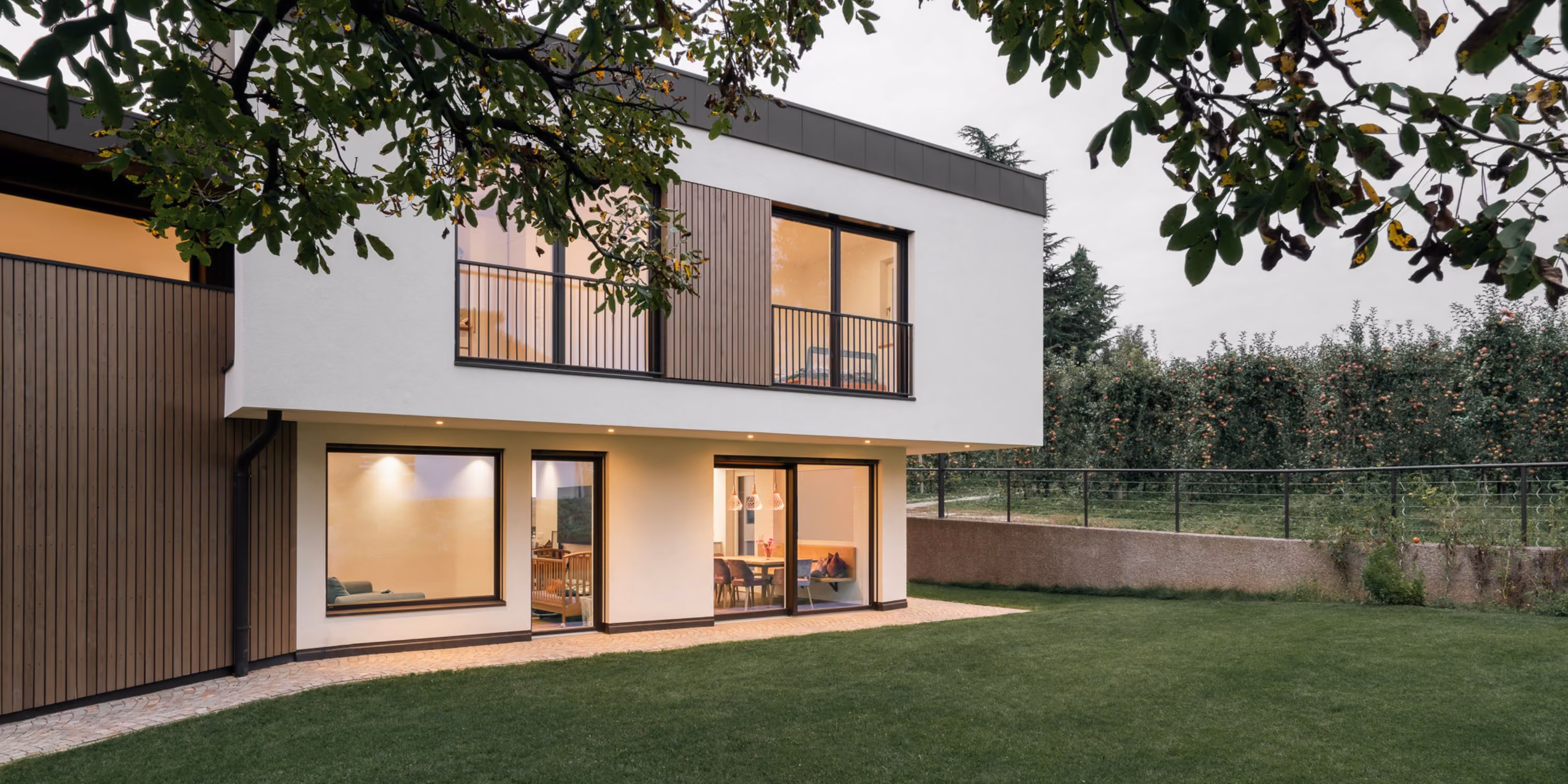 Modern two-story house with white and wooden exterior walls, large windows and doors opening to a green lawn, with a garden and trees in the background under an overcast sky.