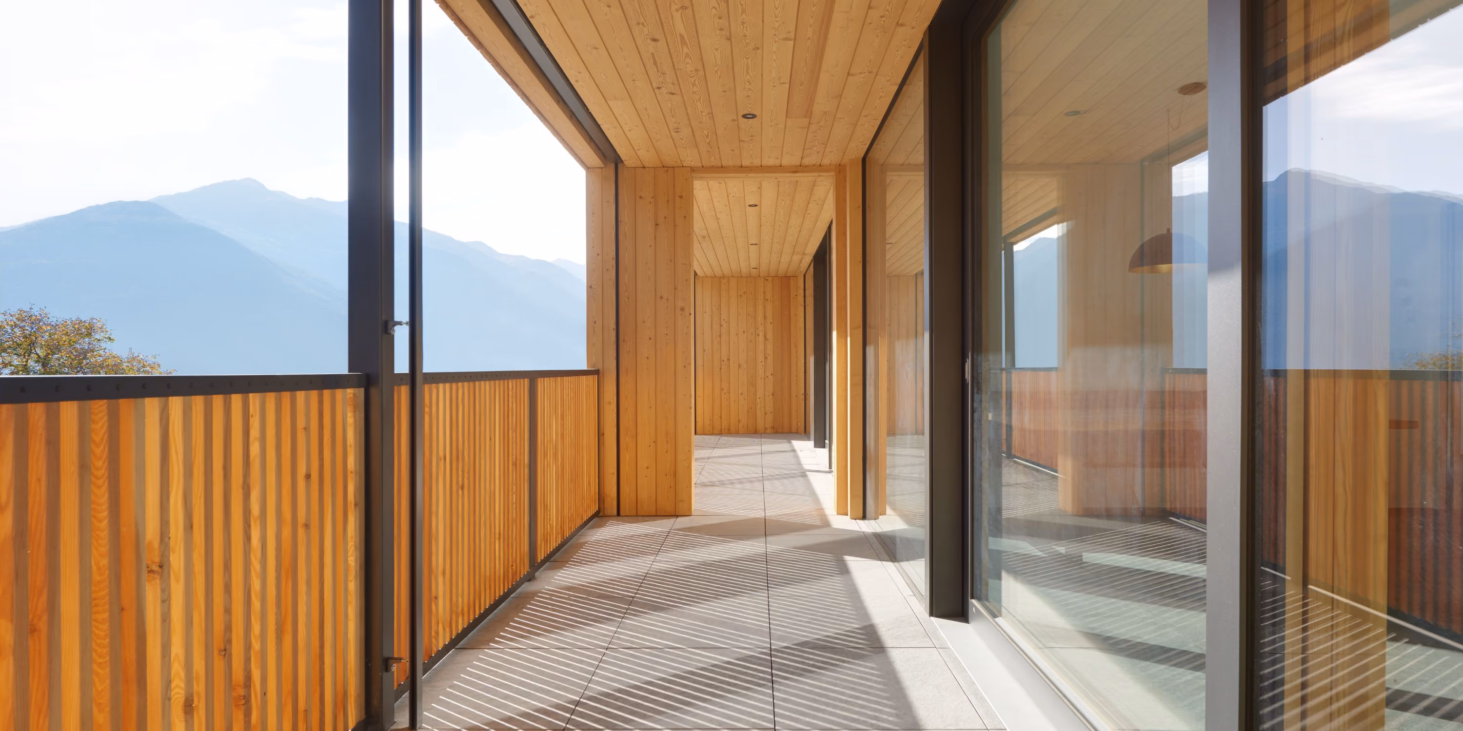 Moderner überdachter Balkon mit Holzverkleidung an Decke und Wänden, Geländer aus Holzlatten und großflächigen Glasfenstern, mit Bergblick im Hintergrund.