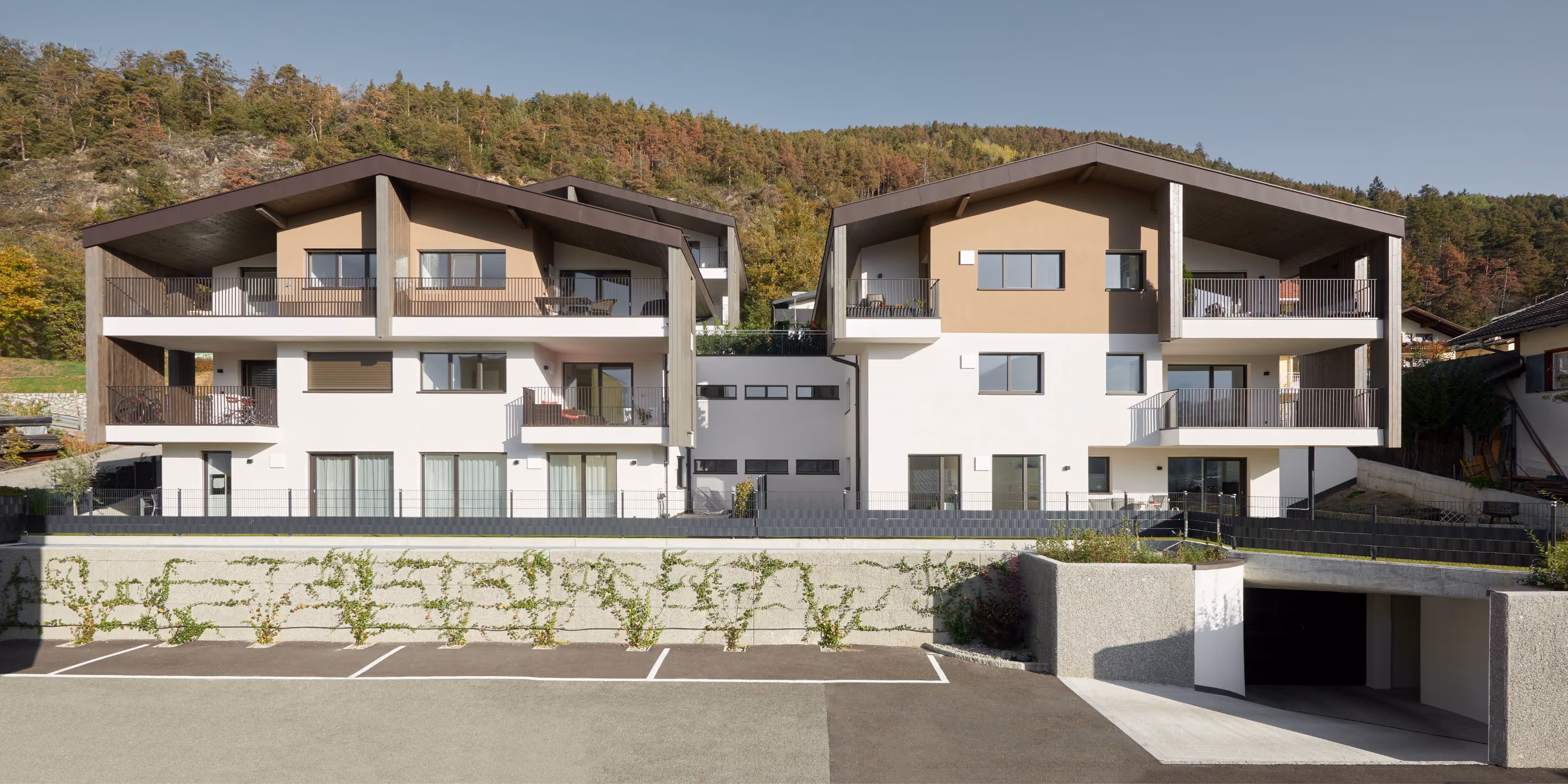 Moderne Wohnanlage mit zwei dreistöckigen Gebäuden, hellen Fassaden mit braunen Akzenten und großen Balkonen, umgeben von grünen Hügeln und Bäumen, davor Parkplätze und Zufahrt zu einer Tiefgarage.