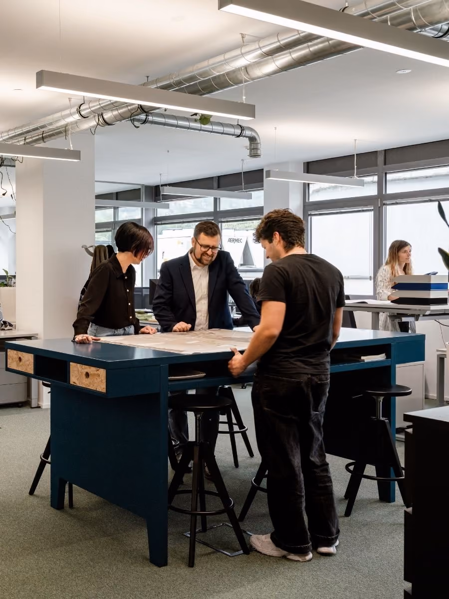 Drei Personen besprechen Pläne auf einem blauen Stehtisch in einem modernen Büro mit großen Fenstern und sichtbaren Lüftungsschläuchen an der Decke, während im Hintergrund eine Frau an einem höhenverstellbaren Schreibtisch arbeitet.