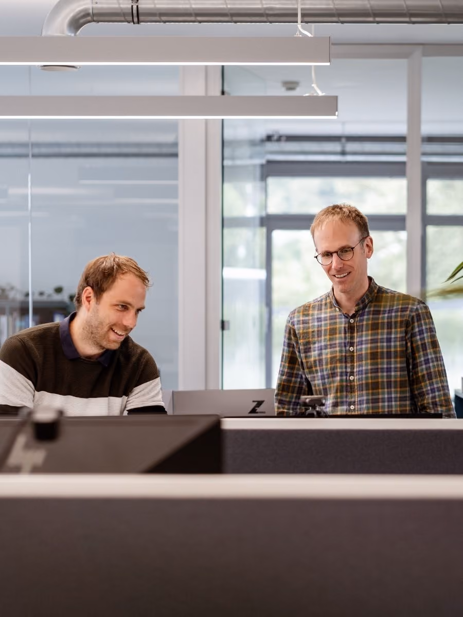 Zwei Männer unterhalten sich lächelnd und arbeiten an einem Laptop in einem modernen Büro mit Glaswänden und Deckenbeleuchtung.