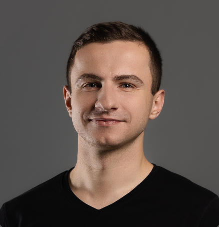 Portrait of a young man with short brown hair wearing a black V-neck shirt against a gray background.