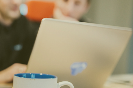 White coffee mug with a blue interior in front of a blurred laptop and two people in conversation.