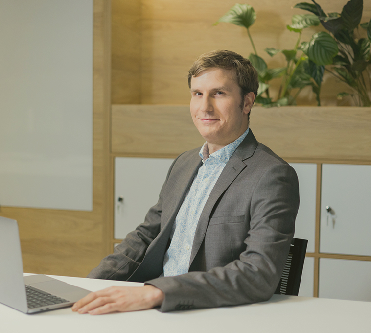 Man in a gray suit sitting at a desk with a laptop in a modern office space with wooden panels and plants.