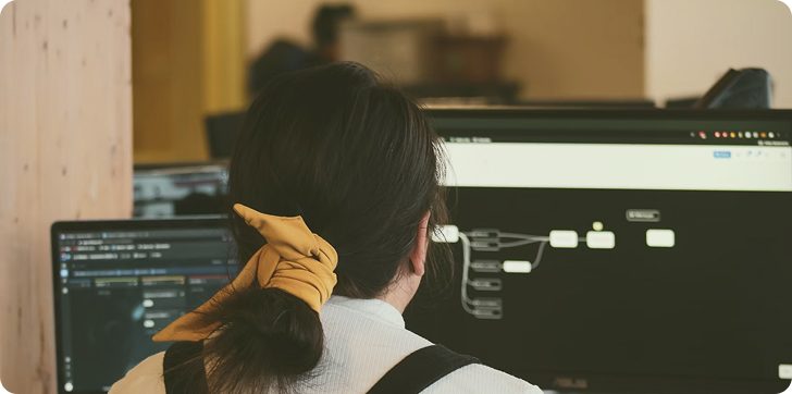 Person with dark hair tied with a yellow scrunchie works on dual monitors displaying technical diagrams and code.