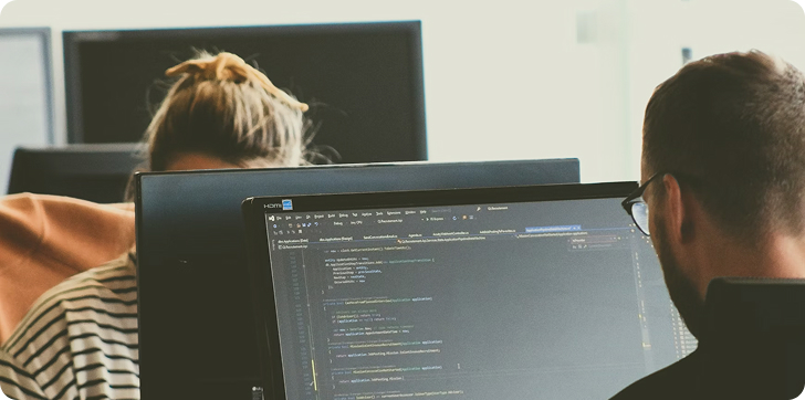 Two people working on computers, one person facing a screen displaying software code.