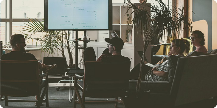 Group of four people sitting in chairs and couches watching a presentation on a screen in a modern office.