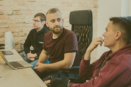 Three men sitting at a desk in an office having a discussion, with laptops and a brick wall background.