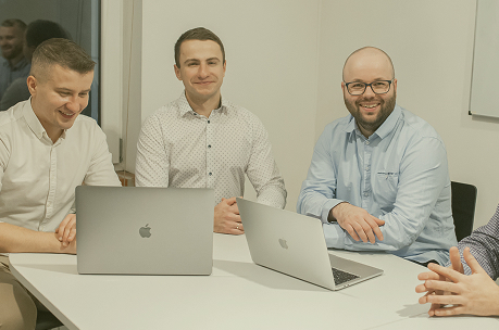 Three men sitting at a table with laptops, smiling and engaged in conversation during a meeting.