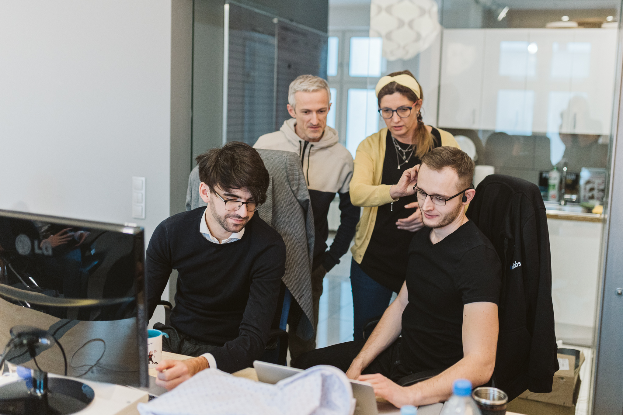 Four colleagues in a modern office collaborating over laptop and desktop computers.