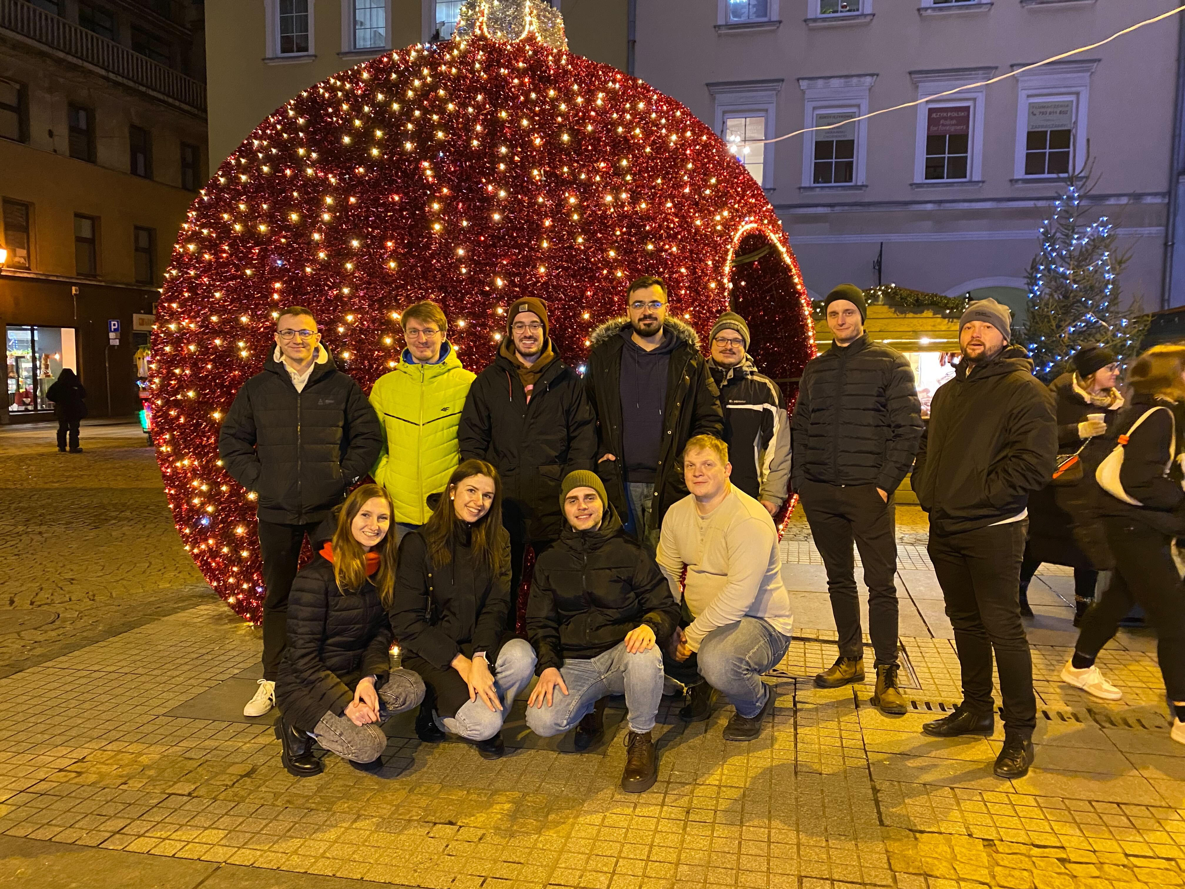 Group of eleven people posing in front of a large, illuminated Christmas ornament decoration on a city street at night.