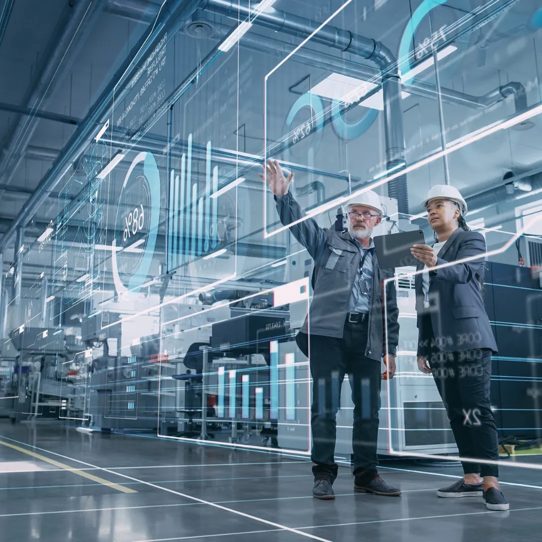 Two engineers with hard hats reviewing digital data charts in a modern industrial factory.
