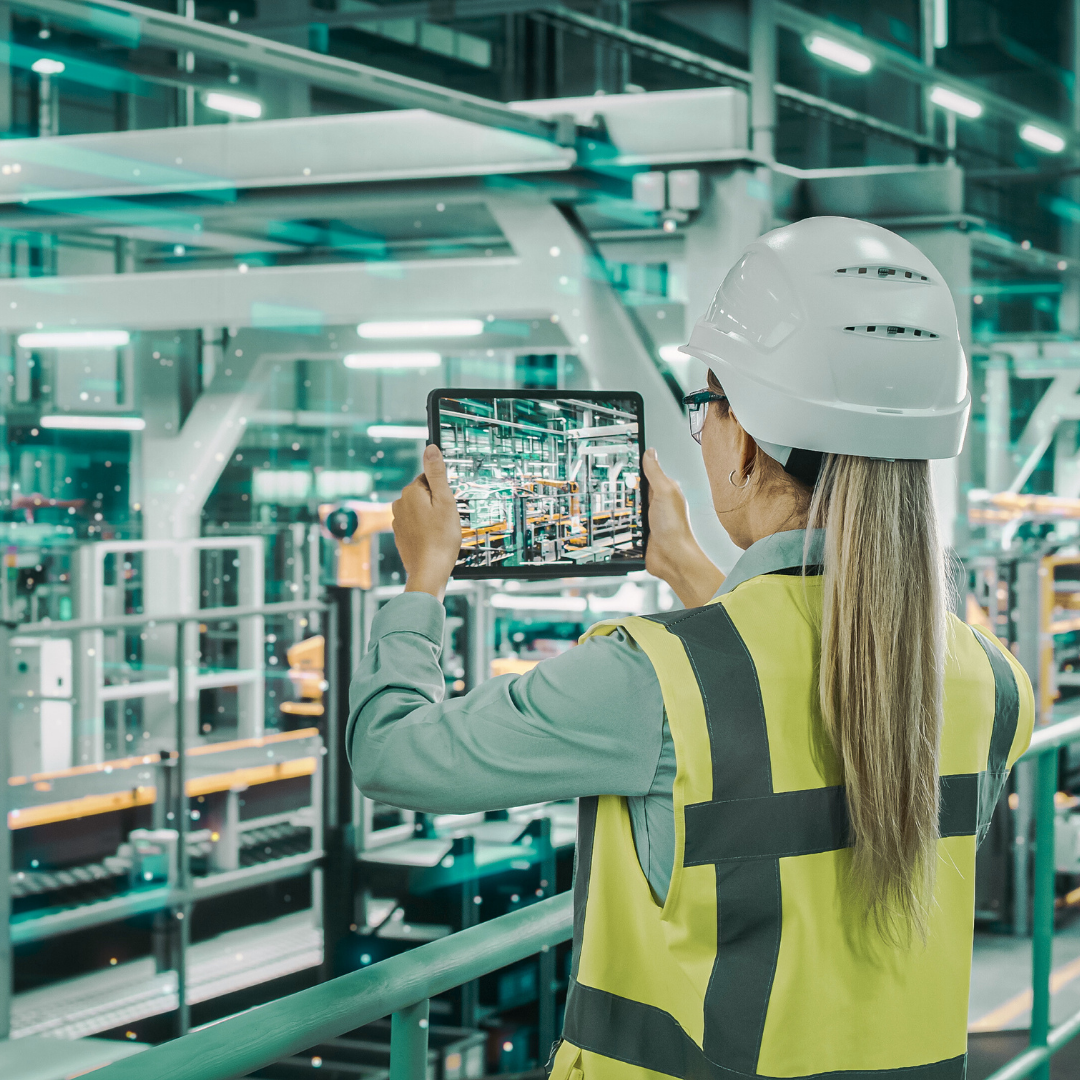 Woman wearing a white hard hat and yellow safety vest using a tablet to inspect an industrial manufacturing facility.