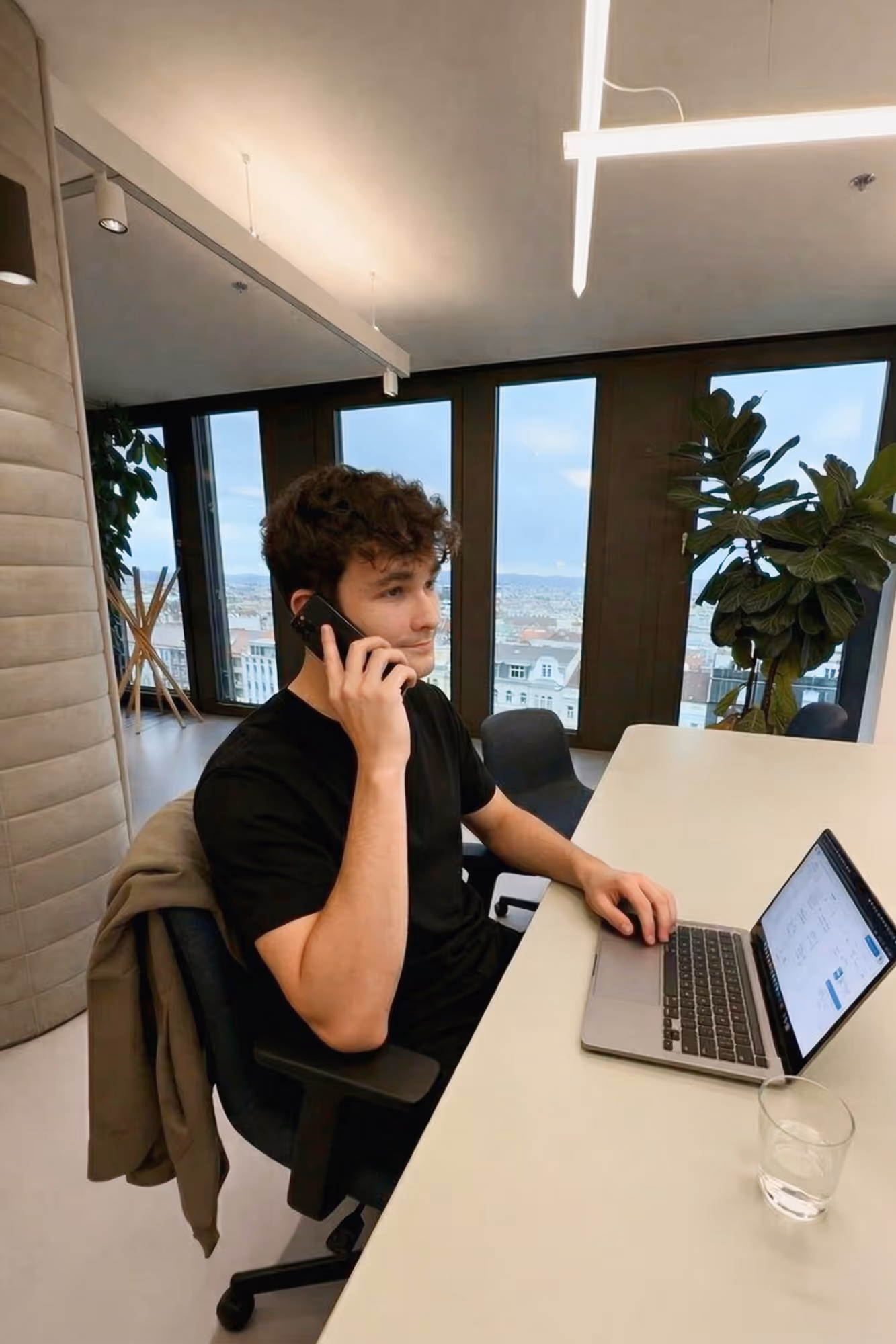 Junger Mann lächelt und telefoniert neben einem Laptop an einem Holztisch in einem hellen Raum.