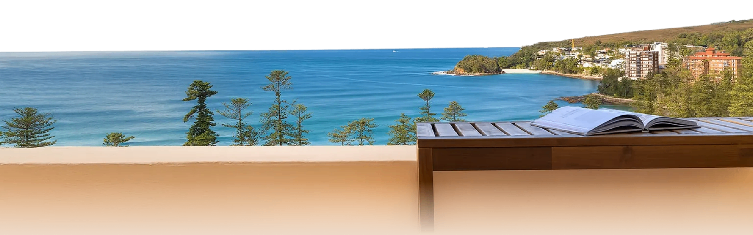 Open book on wooden table overlooking blue ocean, pine trees, sandy beach, and coastal buildings.