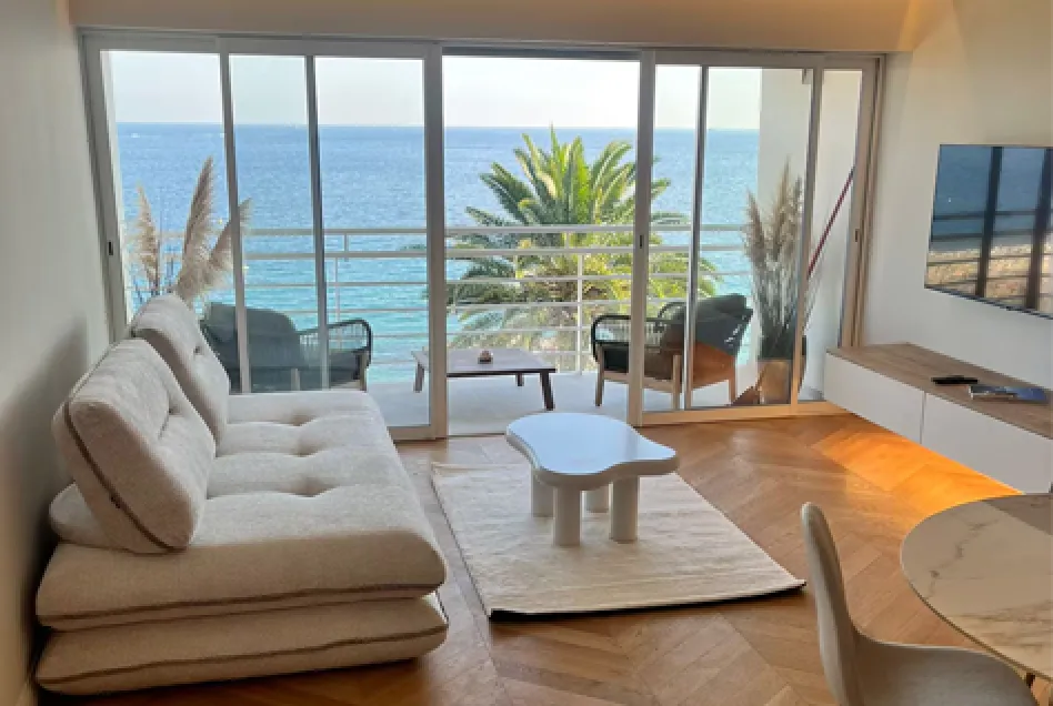 Modern living room with beige sofa, wooden TV stand, sliding glass doors opening to a balcony with ocean view.
