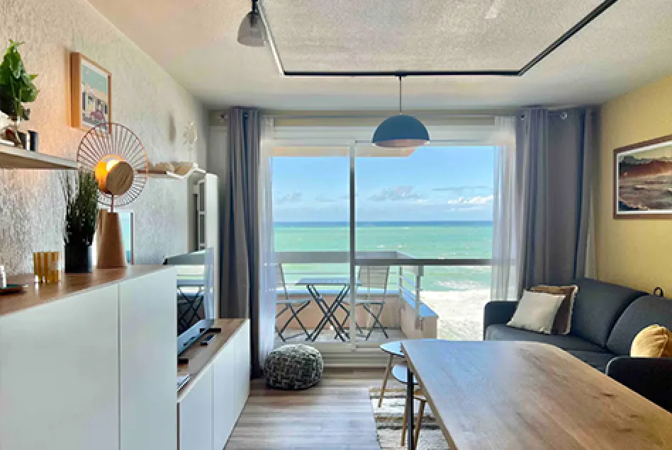 Modern living room with beige sofa, wooden TV stand, sliding glass doors opening to a balcony with ocean view.
