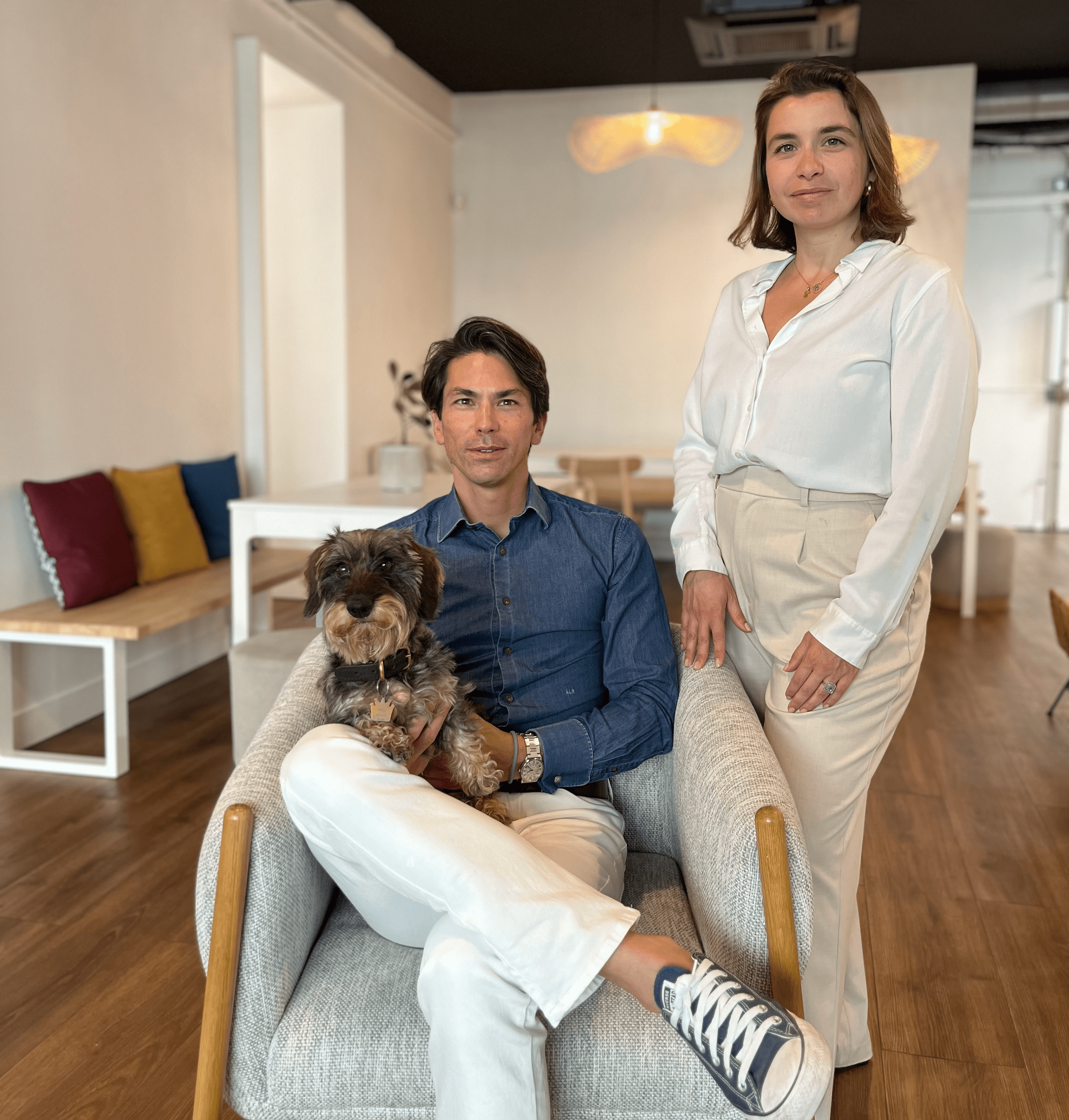 Les deux associés de Comeandwork posent en photo. André est assis sur un fauteuil tenant un teckel sur ses jambes. Fanny est debout à côté du fauteuil.