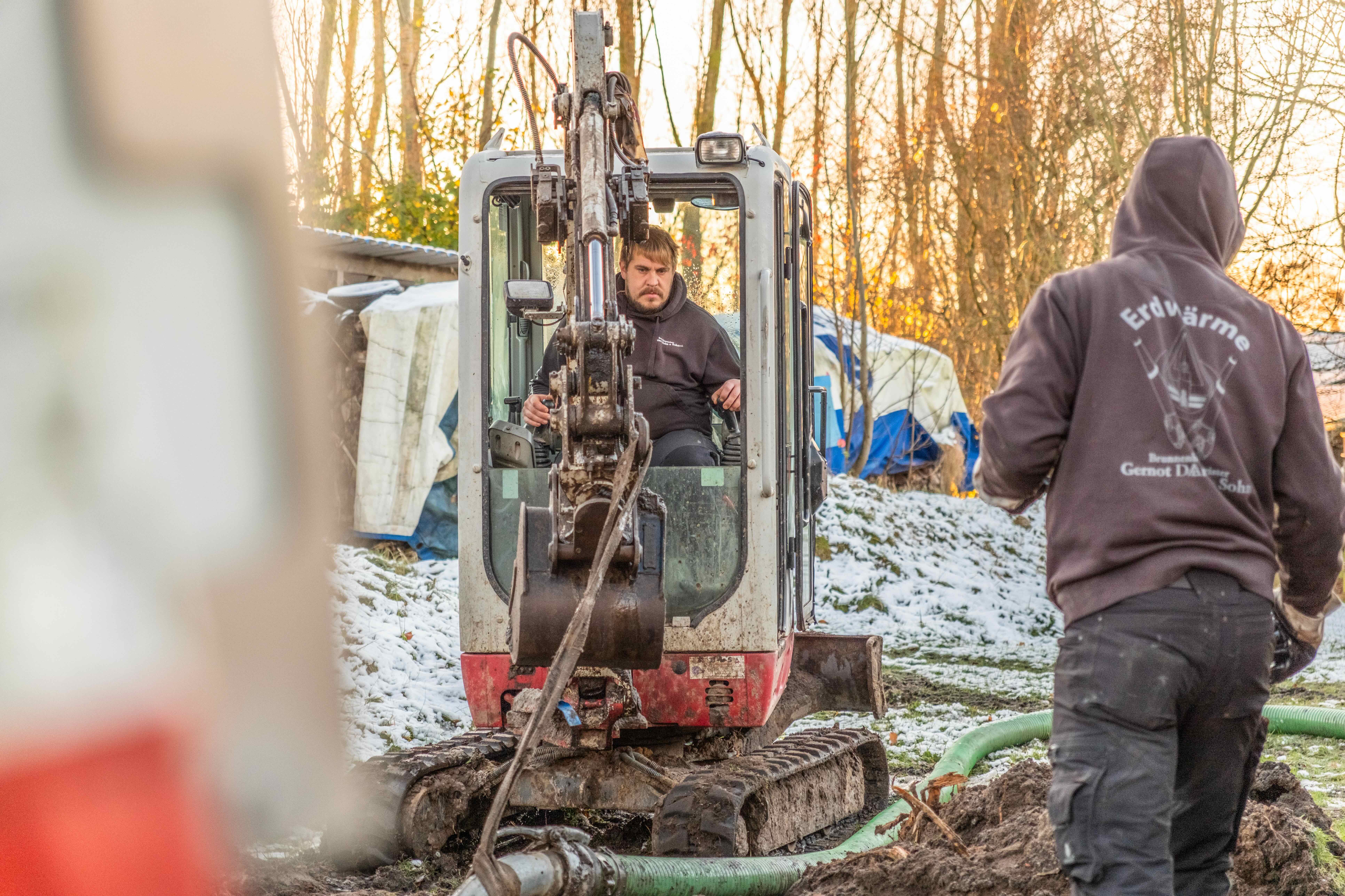 En mand driver en lille gravemaskine, mens en anden mand med en hættetrøje med påskriften 'Geothermal Gernot Daries & Soh' står ved siden af en byggeplads med sne.