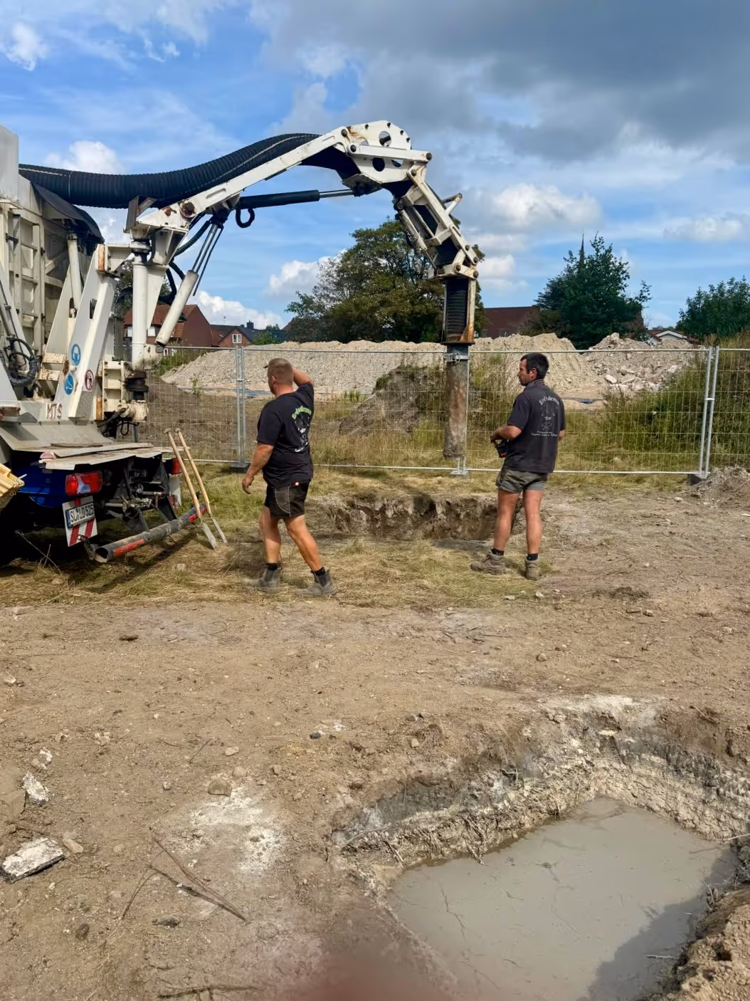 To bygningsarbejdere står ved siden af en lastbil med en stor borerig på en byggeplads med huller i jorden og et indhegnet område i baggrunden.