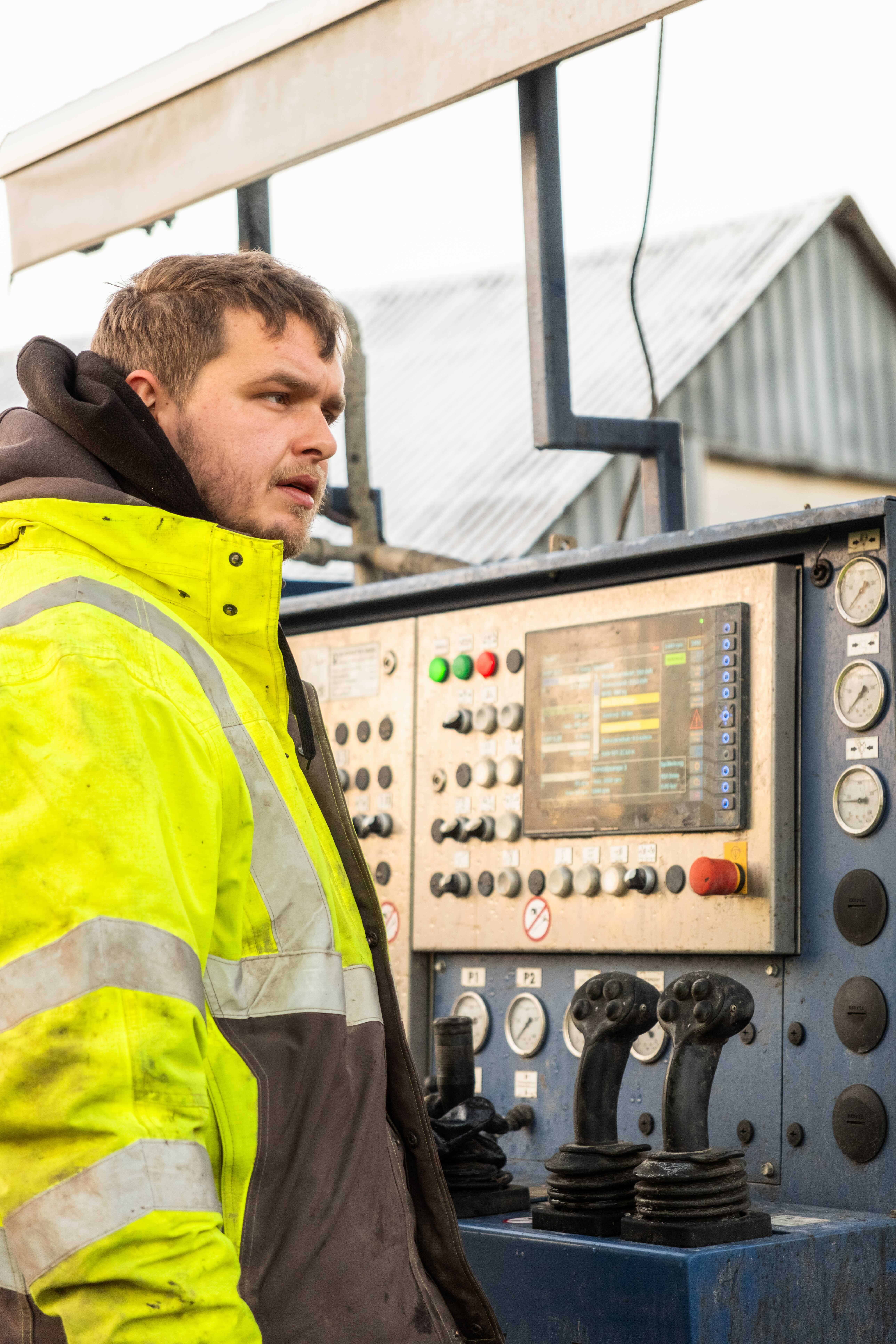 En mand iført en gul advarselsjakke betjener et kontrolpanel med håndtag, knapper og skærme med et koncentreret øje.