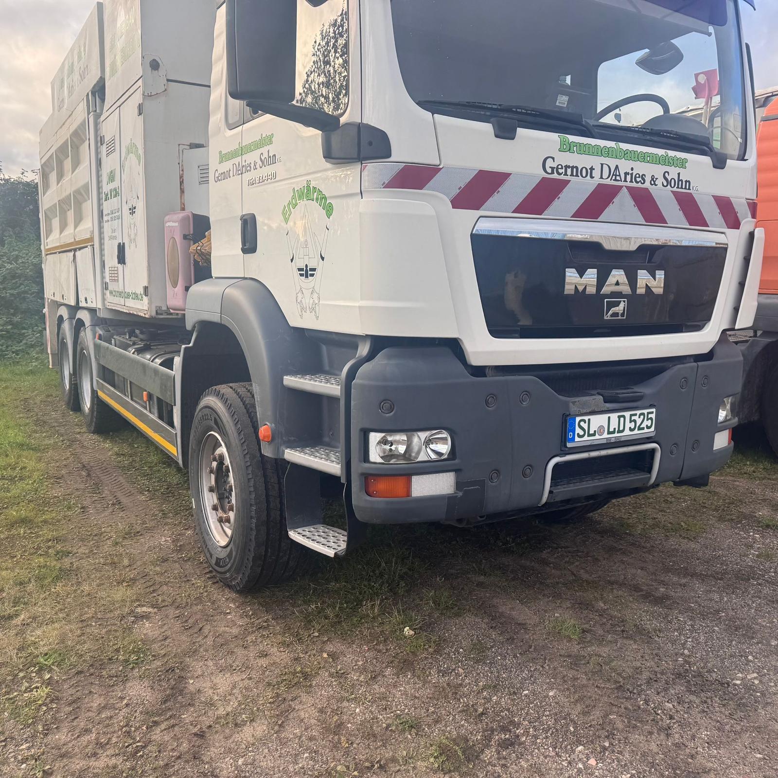 Weißer Saugbagger mit Aufschrift ‚Brunnenbauermeister Gernot DAries & Sohn‘ geparkt auf einem Gras- und Schotteruntergrund.