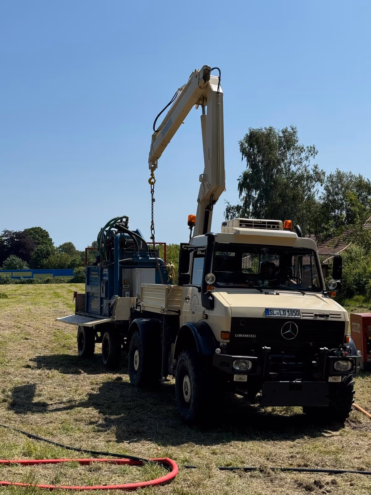 Beiger Unimog-Geländewagen mit Ladekran, der eine Maschine auf einem Anhänger hebt, steht auf einer Wiese bei sonnigem Himmel.