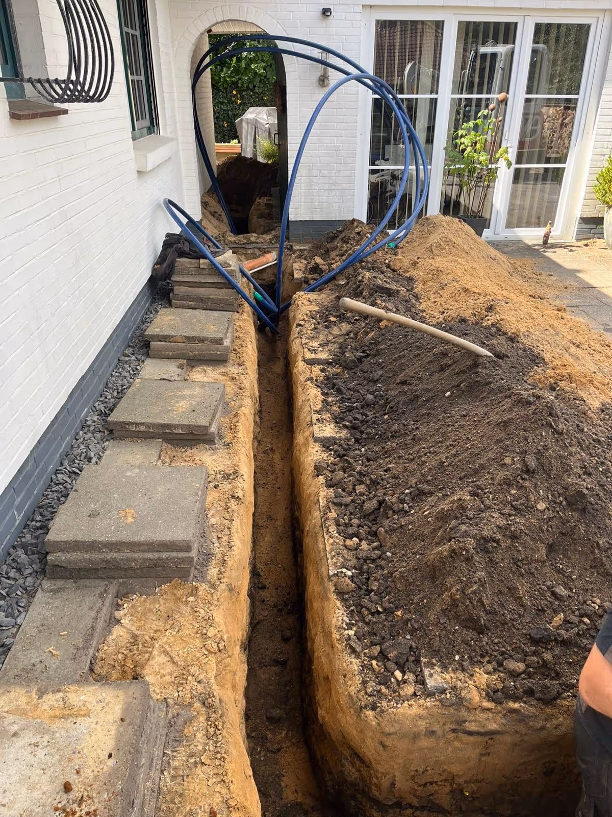Ausgehobener Graben neben einem weißen Haus mit blauen Rohren und aufgetürmtem Erdreich.