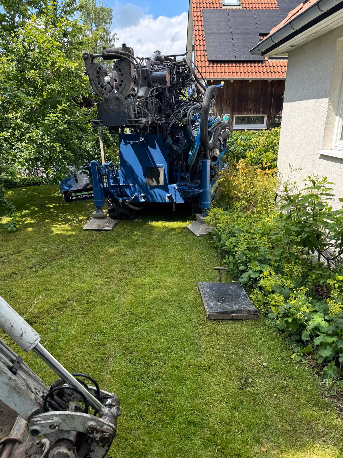 Blaues Bohrgerät steht auf einem gepflegten grünen Rasen neben einem Haus mit Solarzellen auf dem Dach.