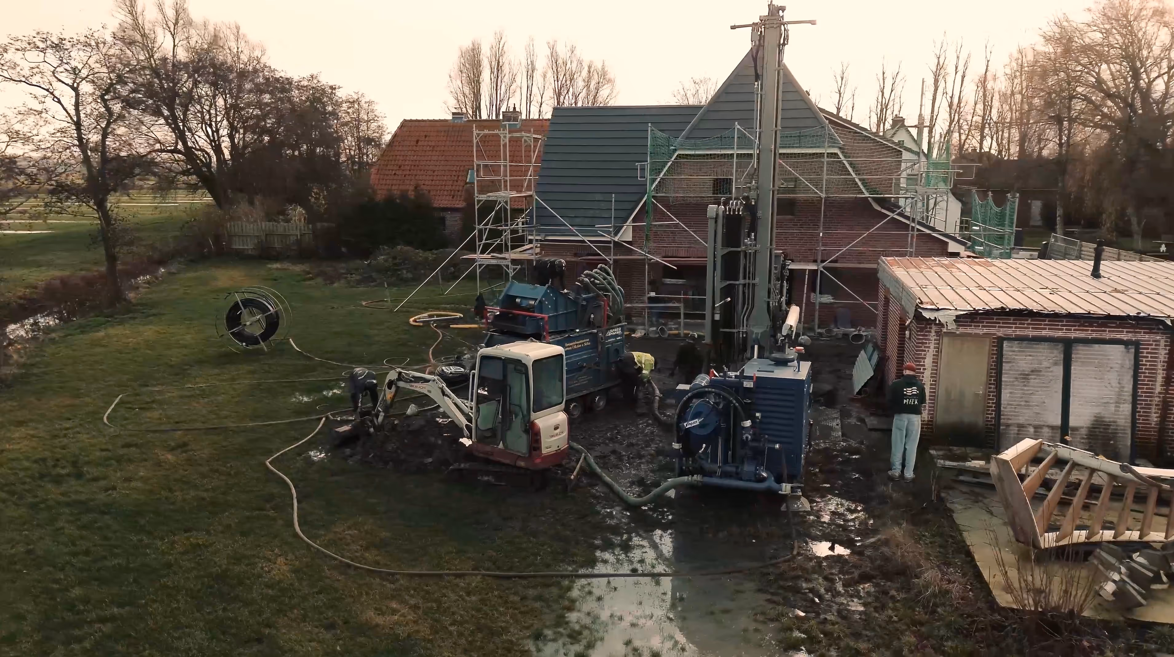 Bauarbeiten mit schweren Maschinen und Arbeitern auf schlammigem Boden vor einem Haus mit Gerüst.