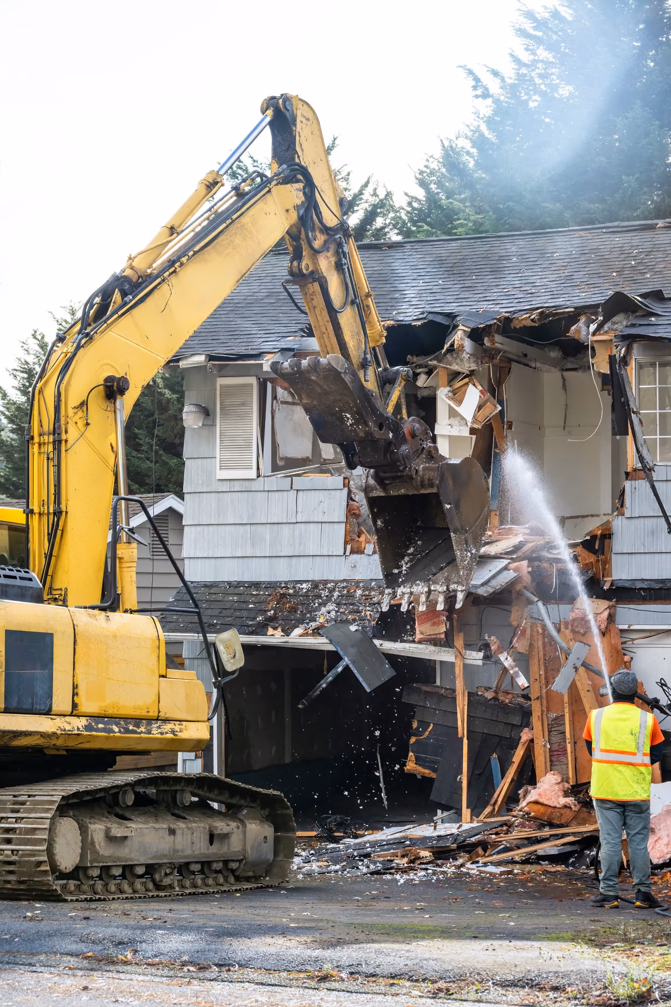 Bagger beim Abbruch eines Hauses, dabei wird die Fassade eines zweistöckigen Gebäudes zerstört, während eine Person in Warnweste und Helm mit einem Wasserstrahl die Staubentwicklung kontrolliert.