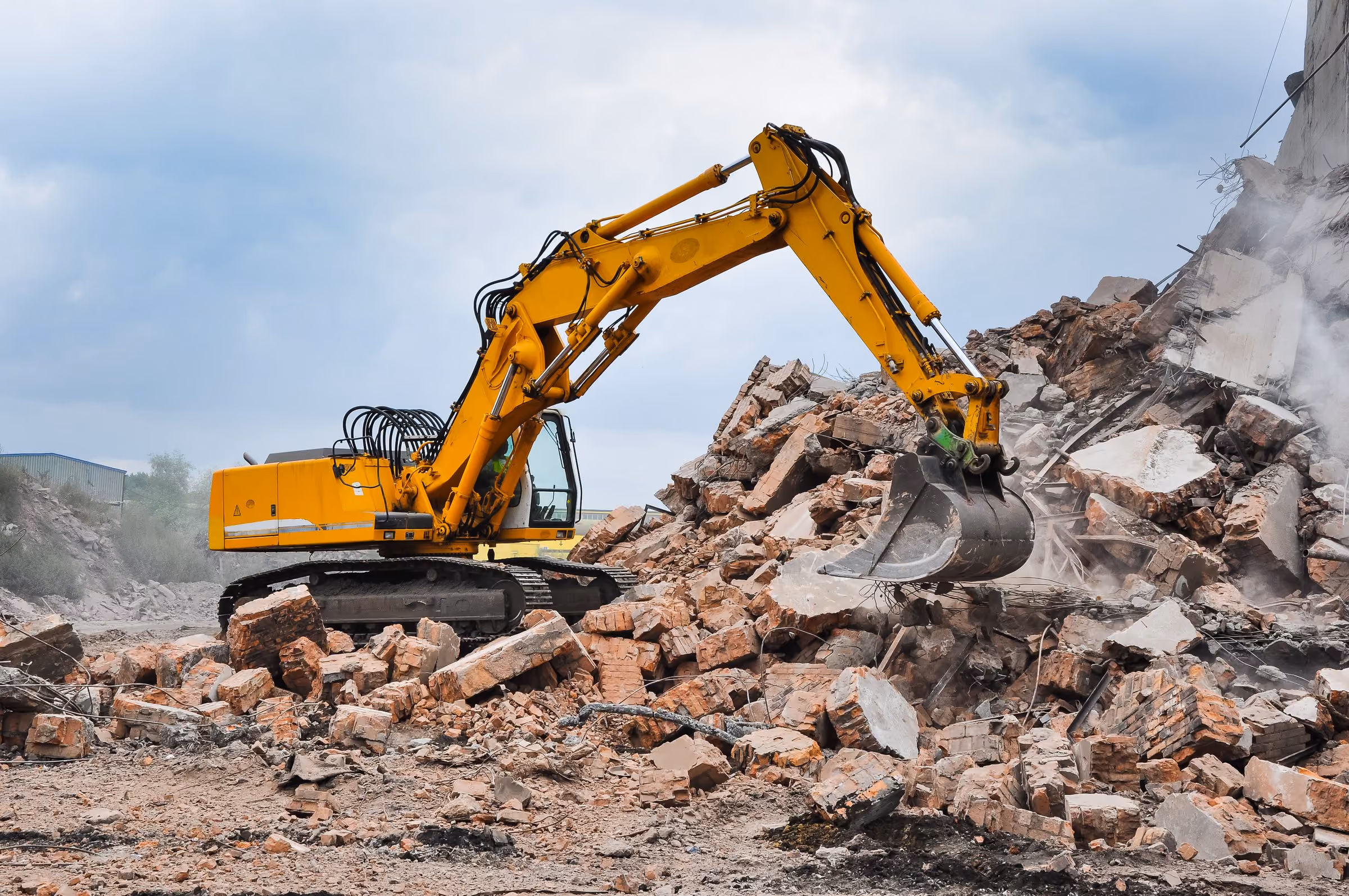 Gelber Abbruchbagger mit langer Ausleger hydraulisch zerlegt Ziegel- und Betonbruchstücke auf einer Baustelle unter bewölktem Himmel.