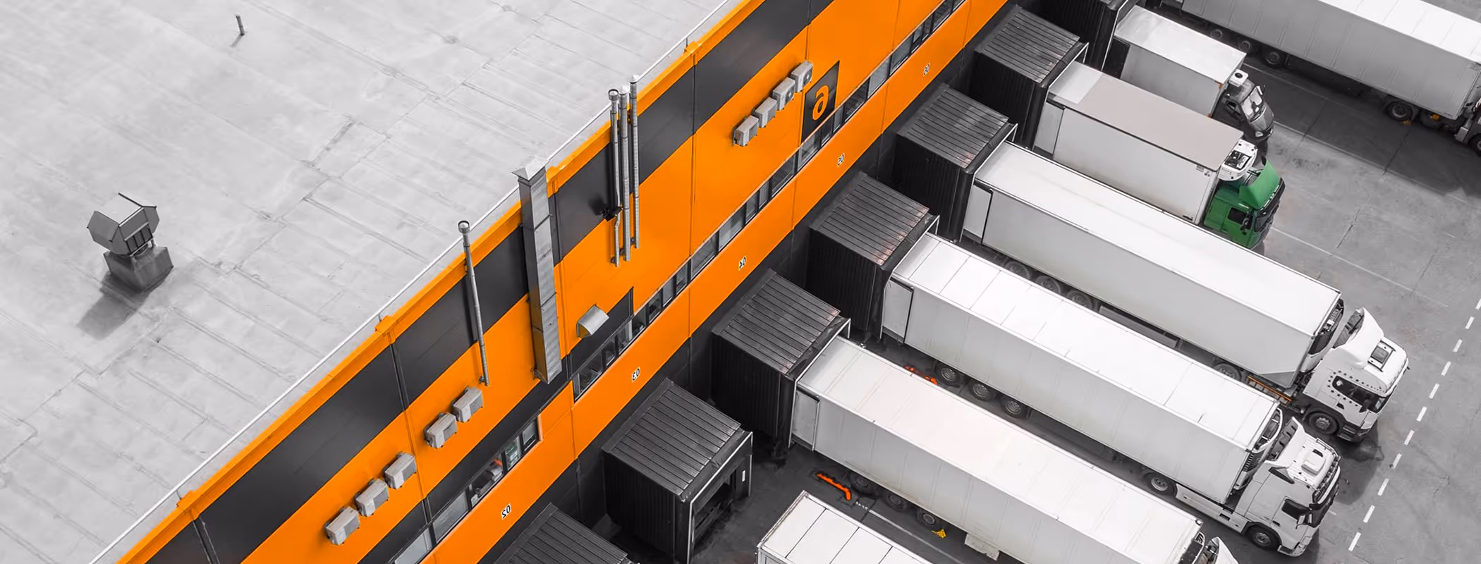Aerial view of multiple white freight trucks docked at loading bays of an orange and black warehouse building.