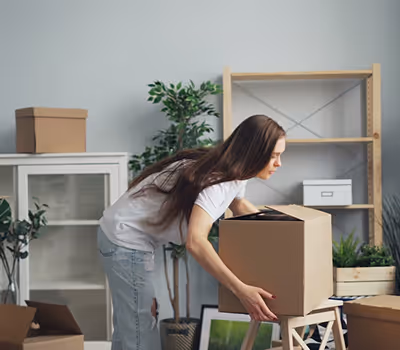 Woman with long brown hair lifting a cardboard box in a room with plants and shelves.
