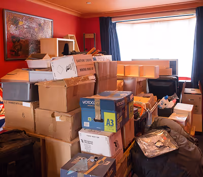 Room filled with stacked cardboard boxes and packed bags near a window with curtains.