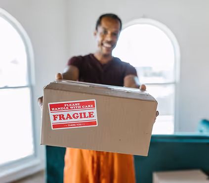 Person holding out a cardboard box with a red label reading 'Fragile, Please Handle With Care'.